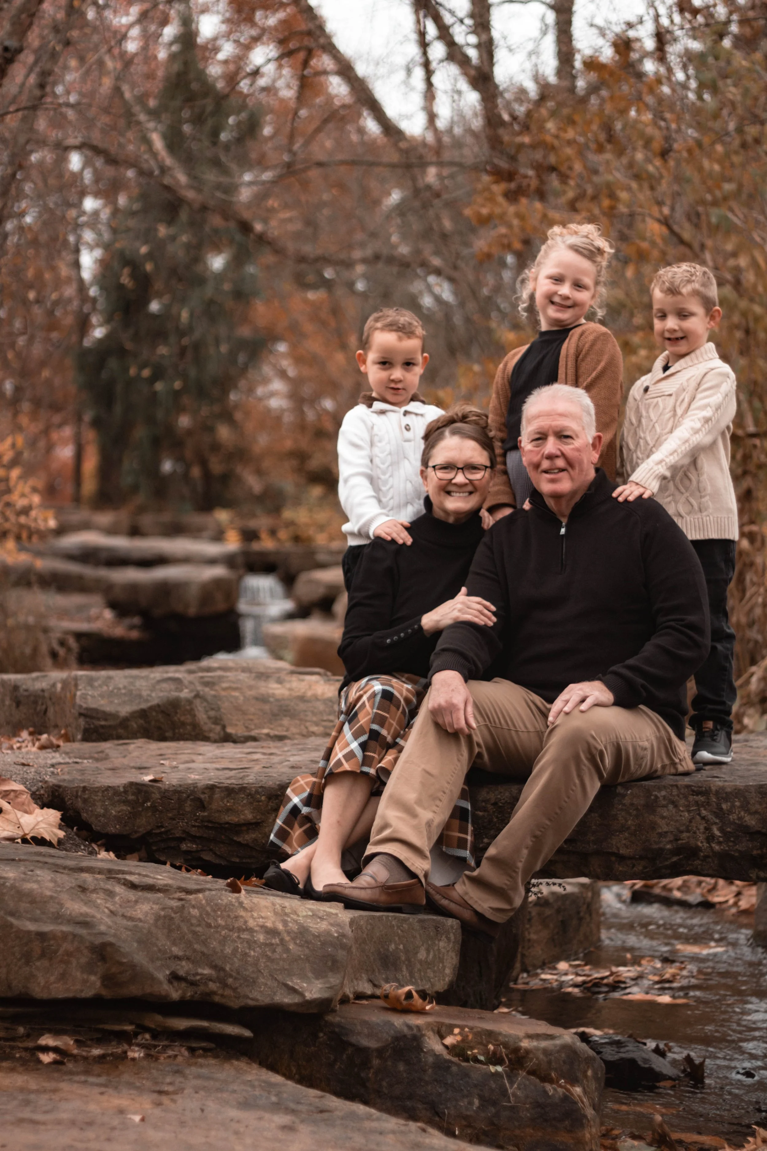 A multi-generational family of six posing outdoors on rocks near a small stream with fall foliage in the background. The family includes two adults seated and four children standing behind them, all smiling.