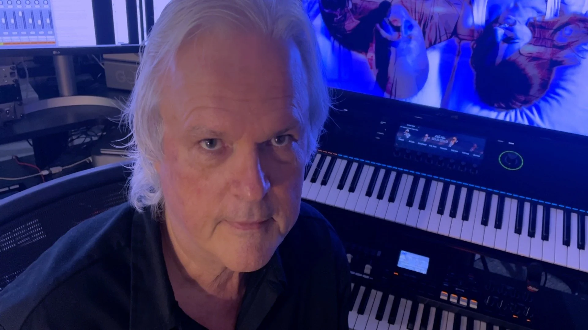 A man with light-colored hair and a serious expression sitting in front of a keyboard synthesizer in a room with music production equipment and a computer monitor displaying digital audio workstations.