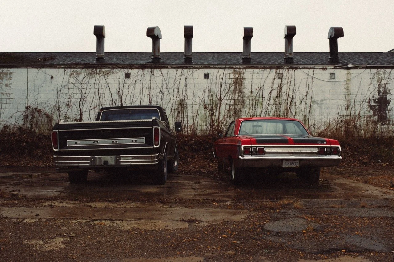 Old black Ford pickup truck and red vintage car parked side by side in a lot, with a weathered white wall and vines in the background.