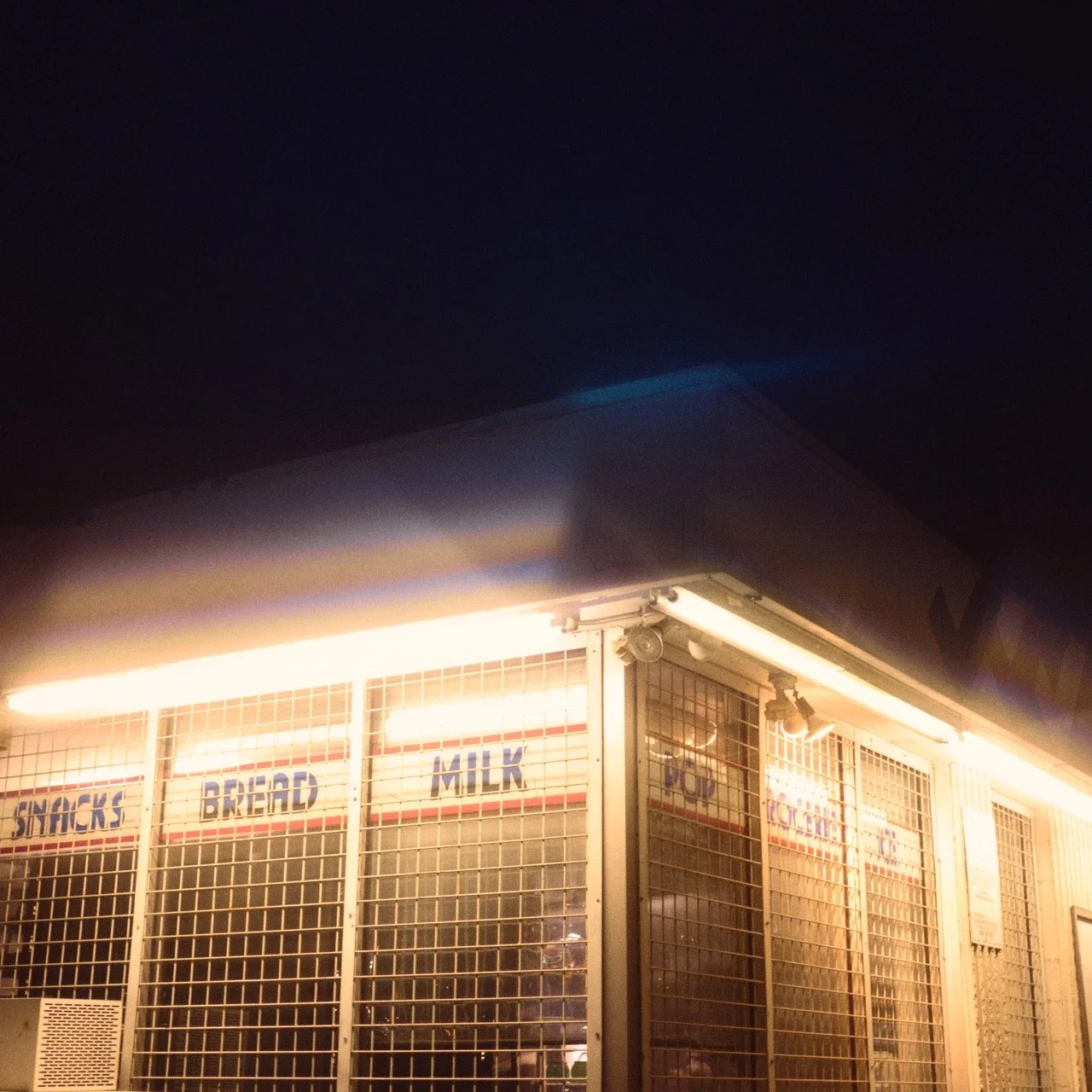 Night view of a store front with illuminated signs reading Snacks, Bread, Milk, and other items, behind metal security grilles.