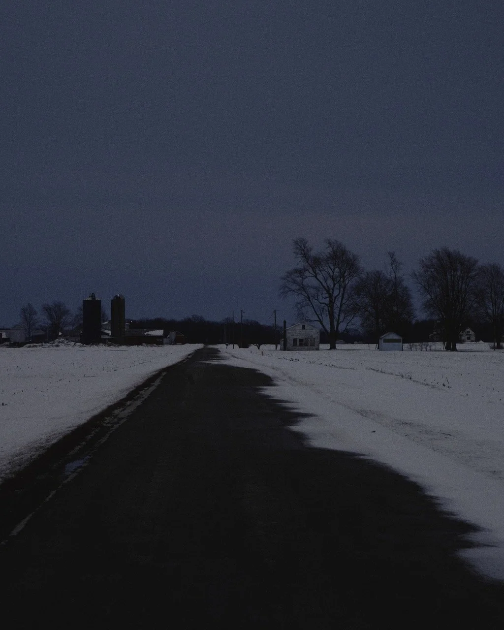 A dark, rural road covered in snow, leading to farm buildings and silos in the distance, with leafless trees and power lines under a moonlit sky.