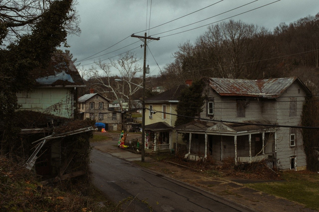 A residential neighborhood with old, worn-down houses, some with broken windows and rusted roofs, on a cloudy day with leafless trees and overhead power lines.