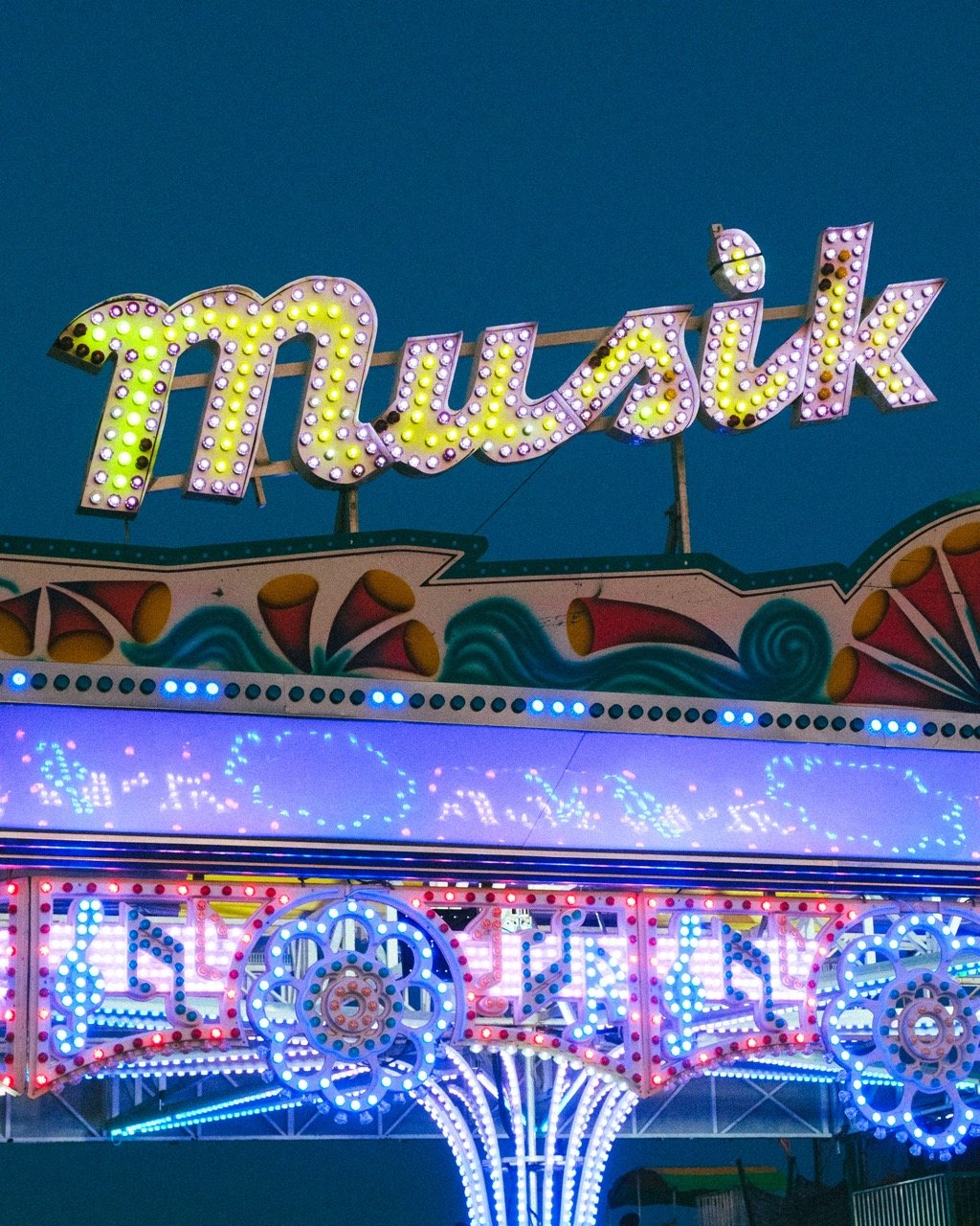 A neon sign at a fairground rides attraction reads 'Moulin Rouge' in illuminated pink and yellow letters against a dark blue sky.