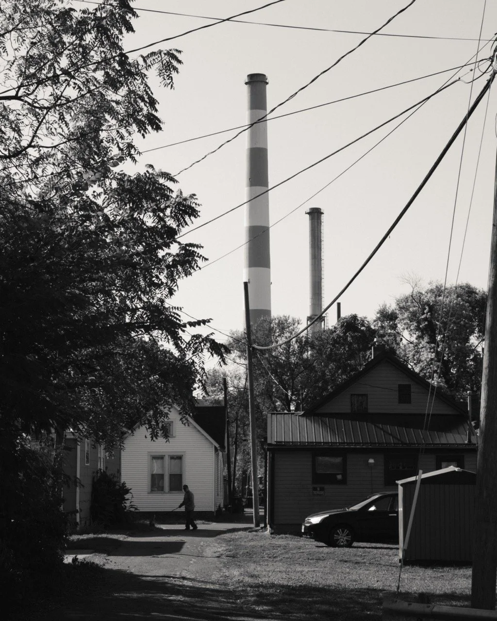 Black and white photo of a residential neighborhood with houses, trees, and a car parked outside. In the background, three industrial smoke stacks are visible.