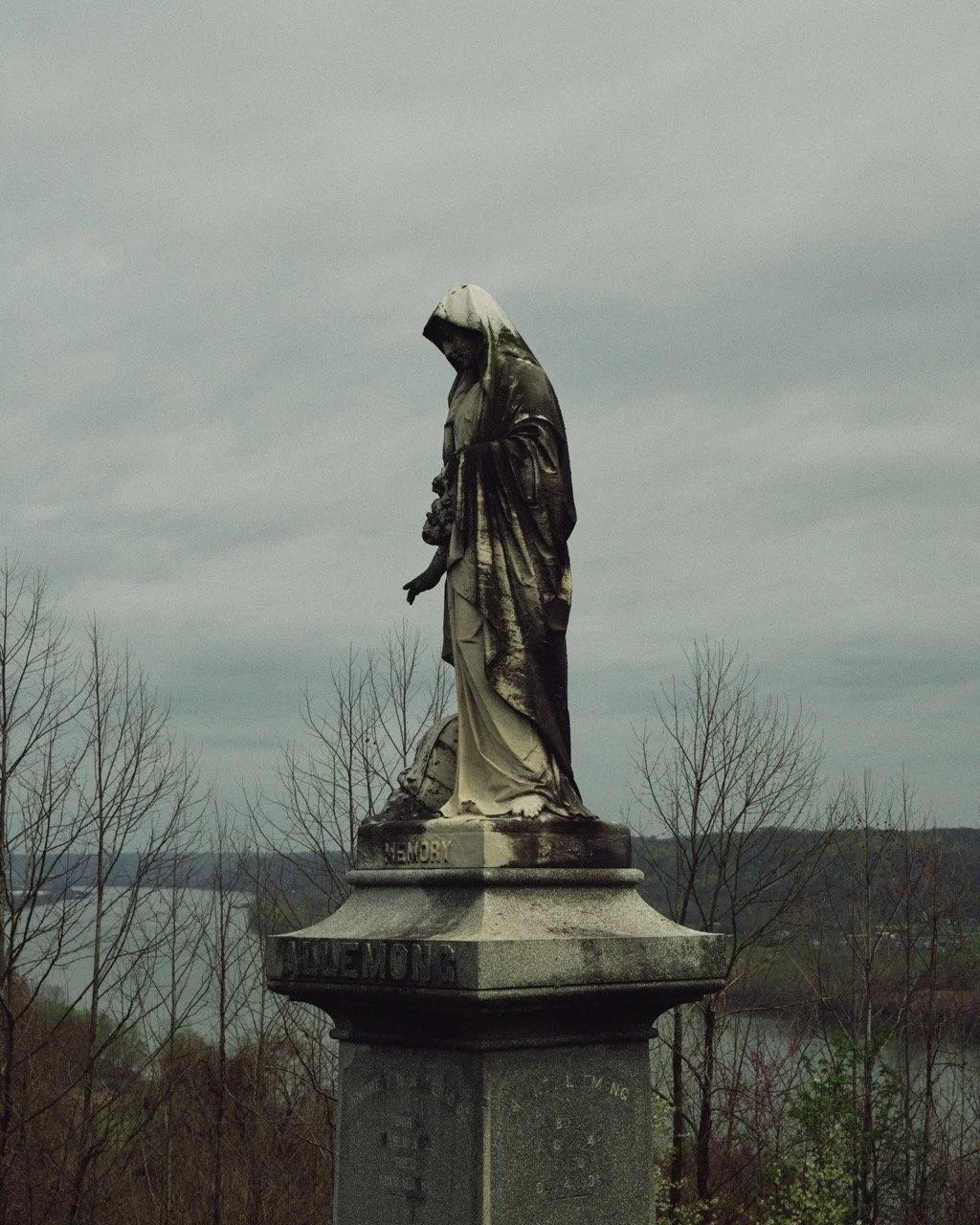 Stone statue of a woman with a hood, standing on a pedestal with trees and water in the background.