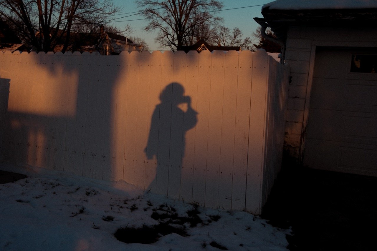 Shadow of a person taking a photo with a smartphone reflected on a white fence during sunset. There is snow on the ground and leafless trees in the background.