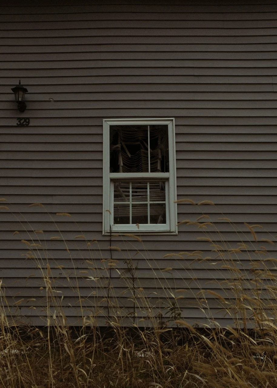 Exterior of a house with gray horizontal siding, a double-hung window, exterior light fixture, and a house number 329. Tall dry grass in the foreground.