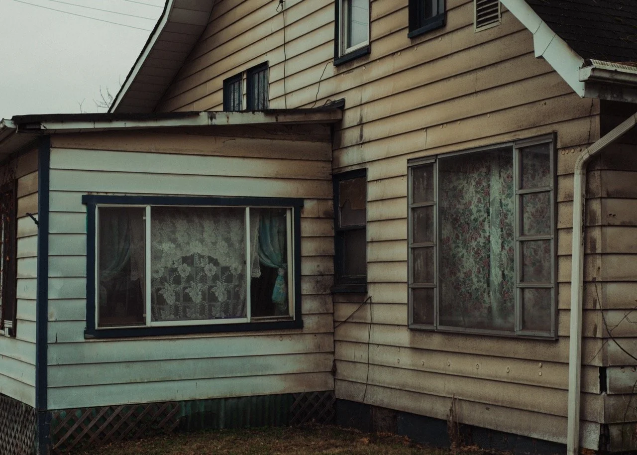Close-up view of an old, weathered house with beige and white siding, dirty windows, and a small, uneven yard.