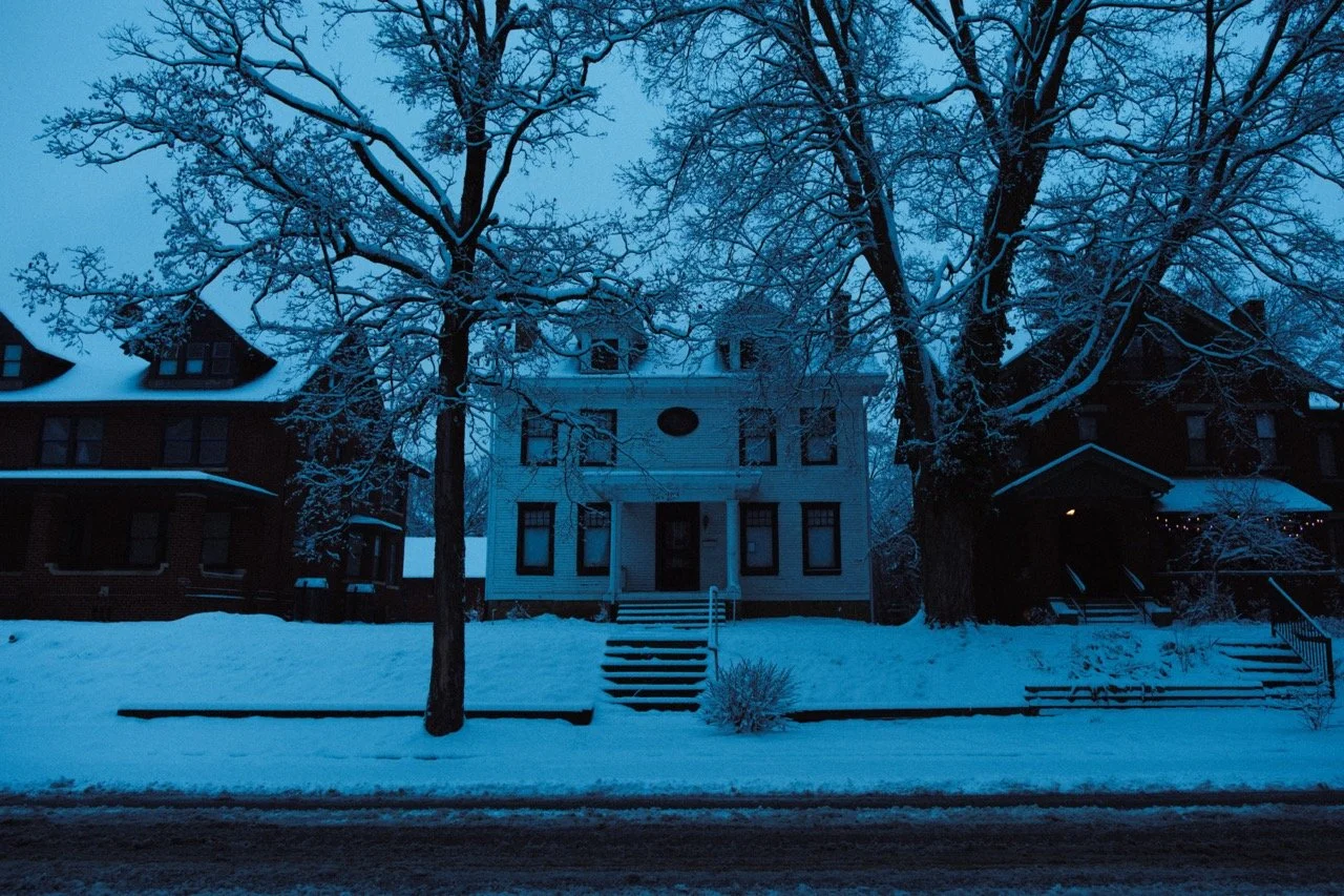 A snowy street scene with a large, white apartment building in the center, flanked by two houses, with snow-covered trees and steps leading up to the buildings.