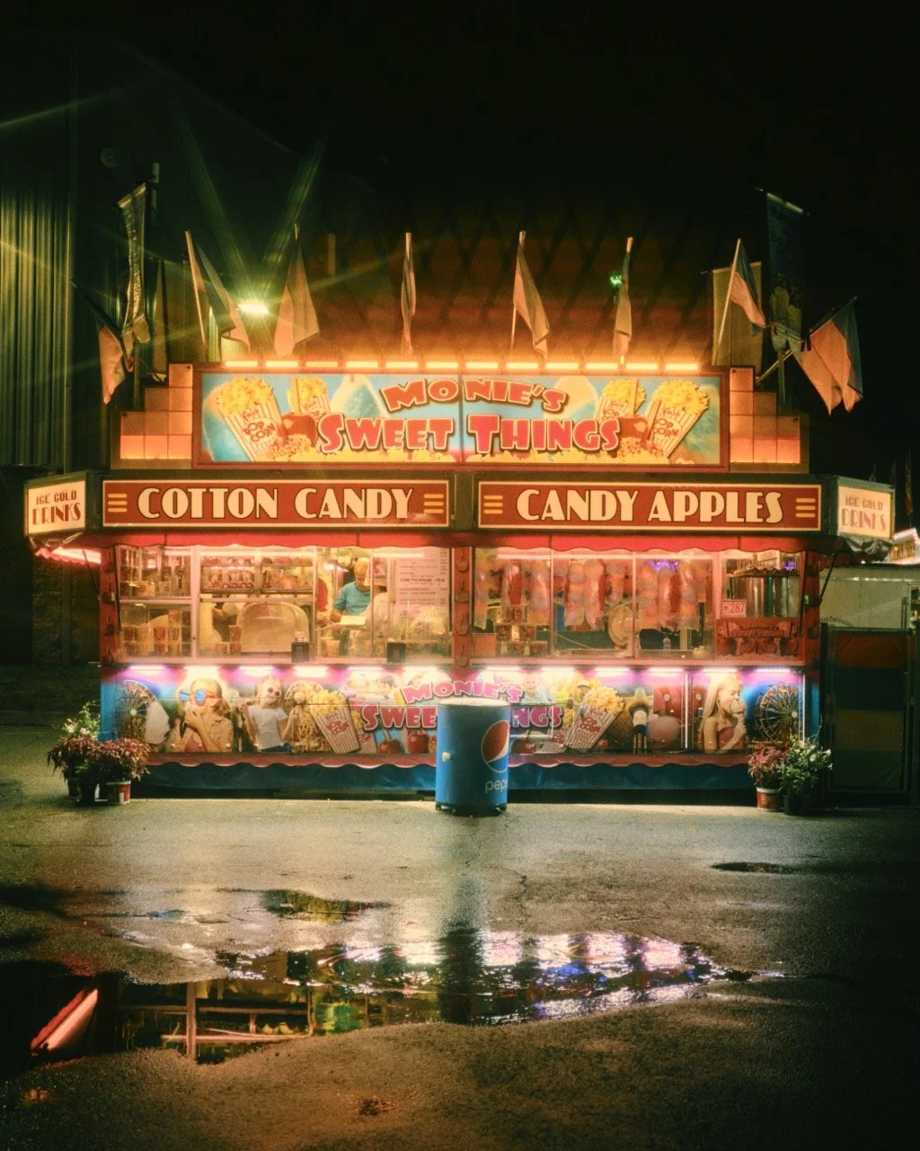 A brightly lit carnival food stand at night selling cotton candy, candy apples, and ice-cold drinks, with colorful signage and flags.