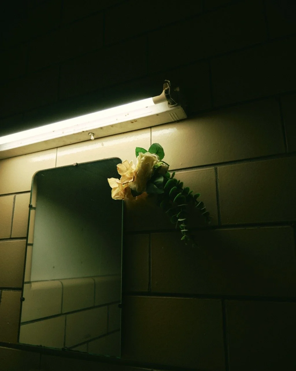 A mirror with a decorative artificial flower and greenery attached to the top, positioned under a fluorescent light fixture on a tiled wall.