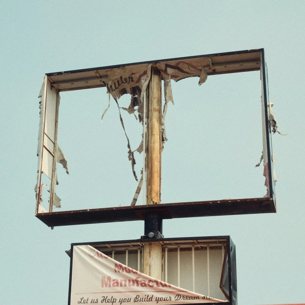 A damaged, empty billboard frame with torn remaining fabric, against a clear blue sky.