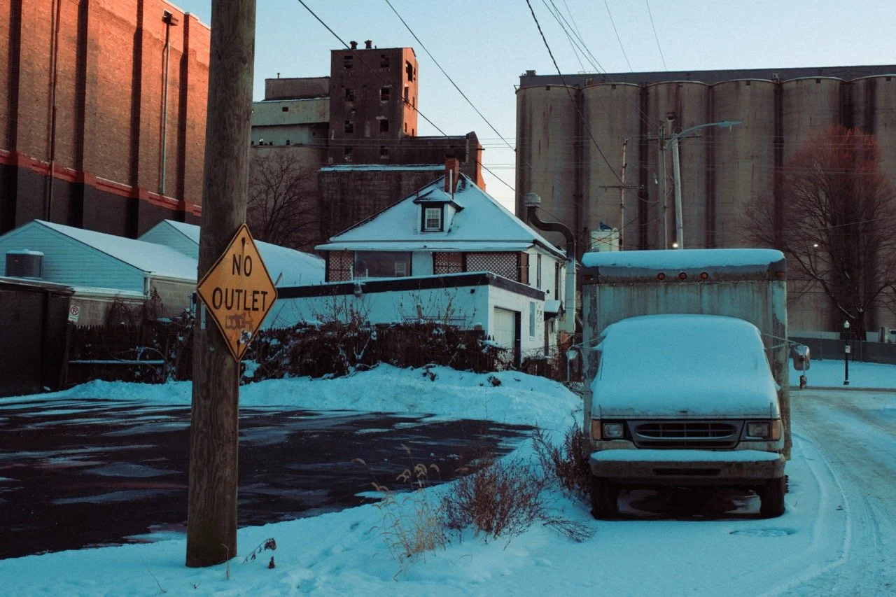 A quiet street scene in winter with snow covering the ground and vehicles. There is a white house with a gabled roof, a large No Outlet sign on a utility pole, a snow-covered delivery truck, and a dark asphalt street with some patches of snow and ice