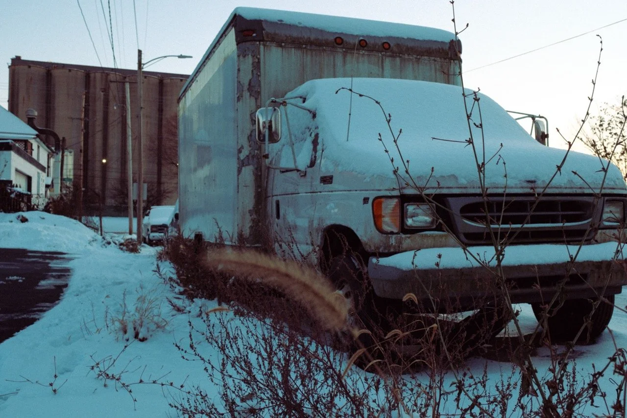 An old, weathered delivery truck covered in snow parked on a snowy residential street with houses, power lines, and a fence in the background.