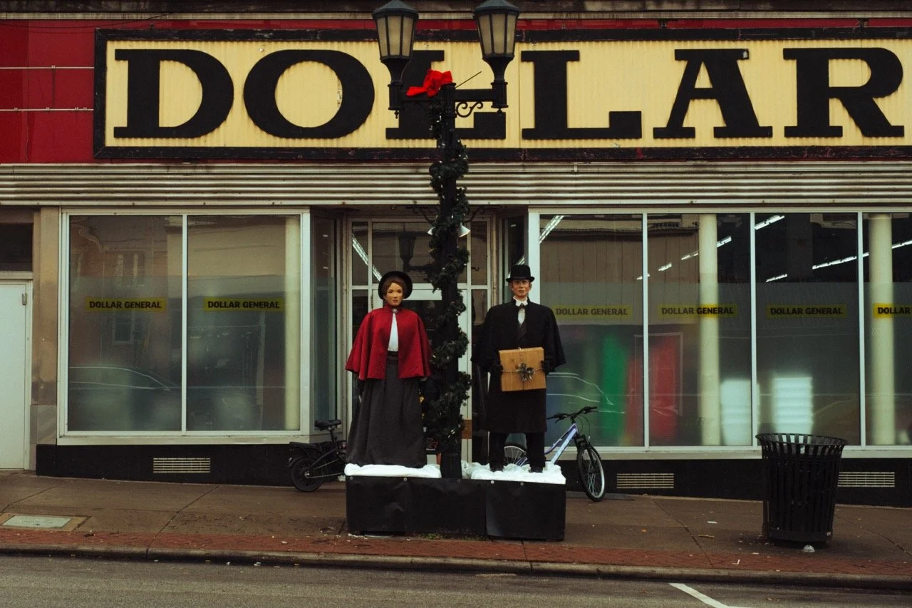 Empty storefront with a large sign reading 'DOLLAR'. Two mannequins dressed as holiday figures are in front, one with a red cape and the other holding a gift box. A lamppost decorated with Christmas greenery and a red bow is between the mannequins. T