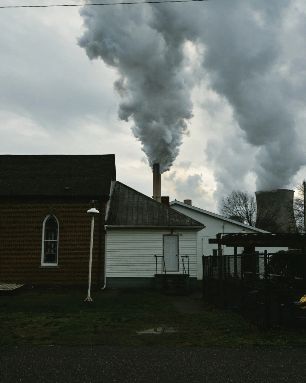 Industrial area with two smokestacks emitting thick smoke into the cloudy sky, adjacent to residential buildings.