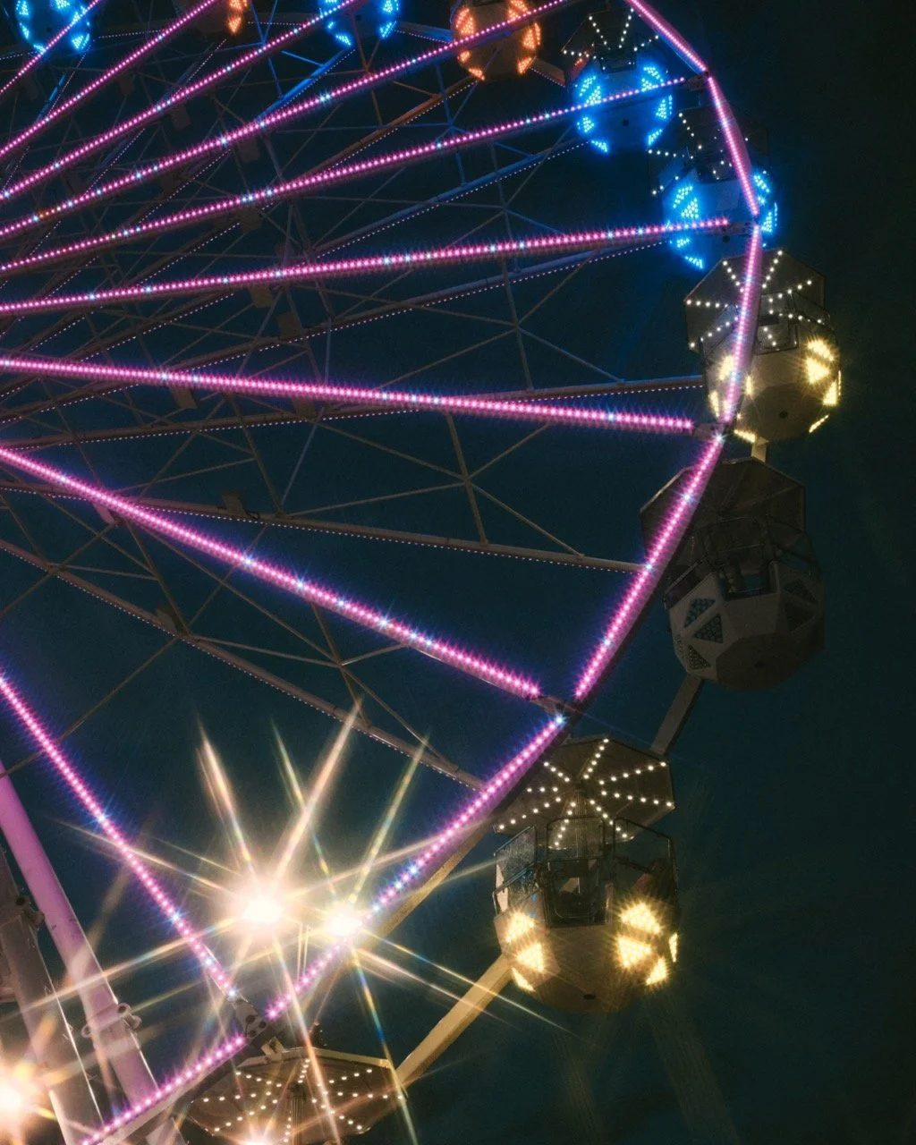 A brightly lit Ferris wheel at night with colorful pink, blue, and yellow lights and illuminated cabins against a dark sky.