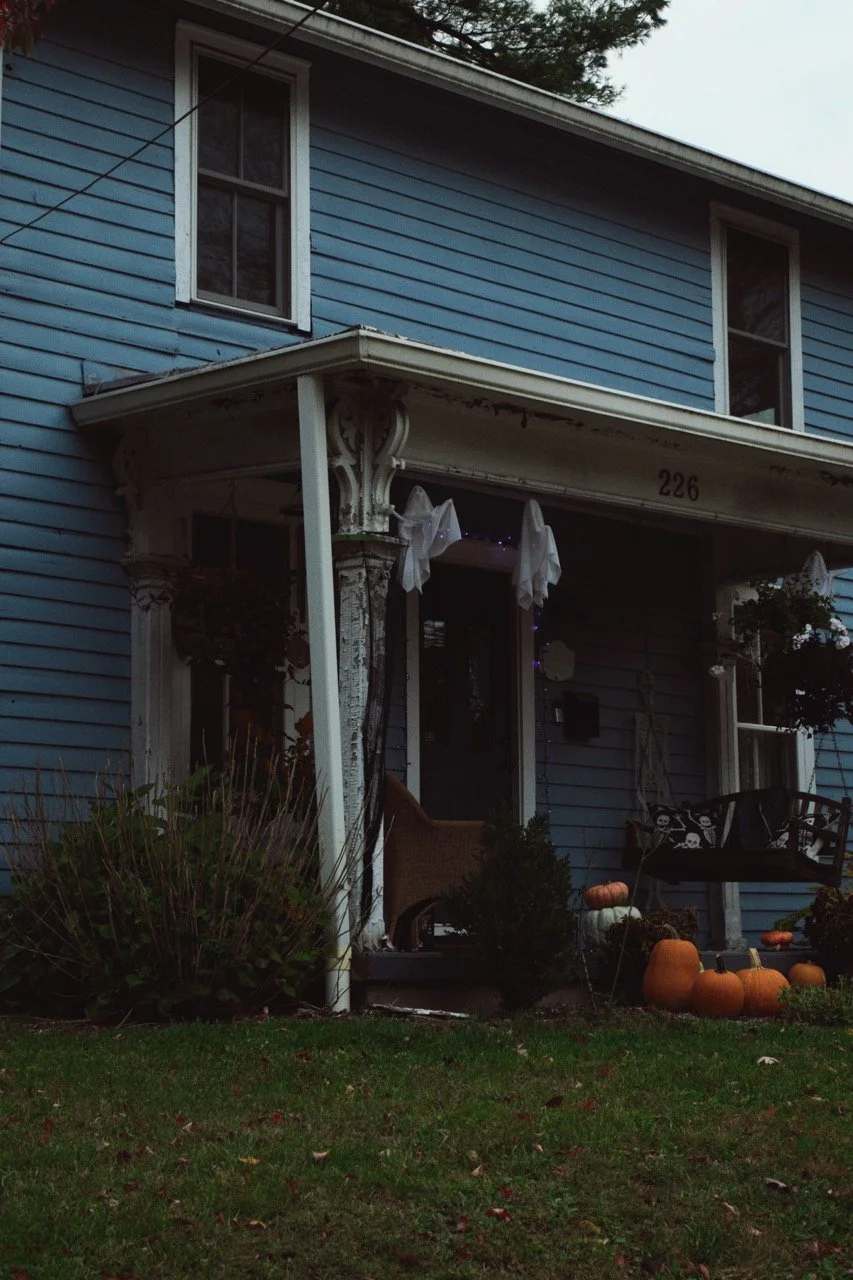 A blue two-story house with a porch decorated for Halloween and fall. There are pumpkins, a bench, hanging flowers, and Halloween-themed decorations near the entrance.