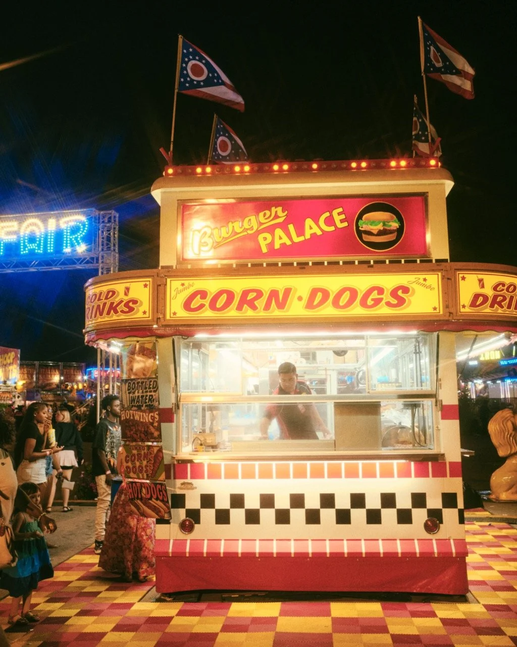 Night carnival scene with a hot dog stand named 'Burger Palace' decorated with Ohio flags on top. Several children and adults are waiting or walking nearby, with bright lights and amusement rides visible in the background.