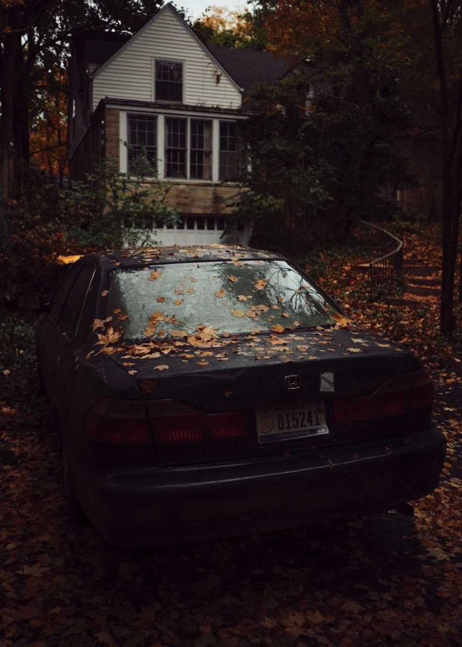 A black Honda car parked in front of a white house with a porch, surrounded by trees and fallen autumn leaves.