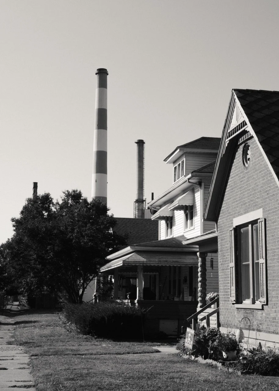 Black and white photo of a residential neighborhood with houses, trees, and smoke stacks in the background.