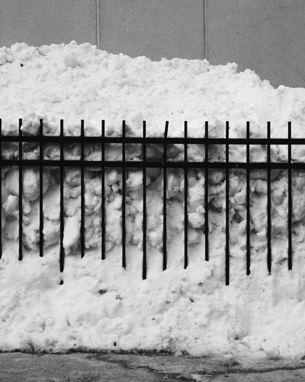 A large pile of snow behind a black metal fence, with a plain wall in the background.