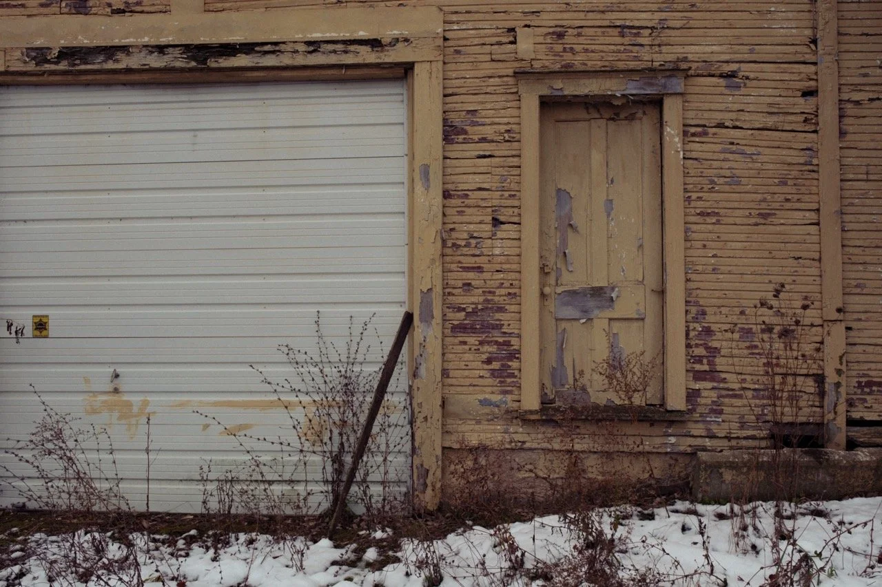 An old, abandoned building with a closed garage door and boarded-up window. The building's paint is peeling, and there are dried plants and snow on the ground.