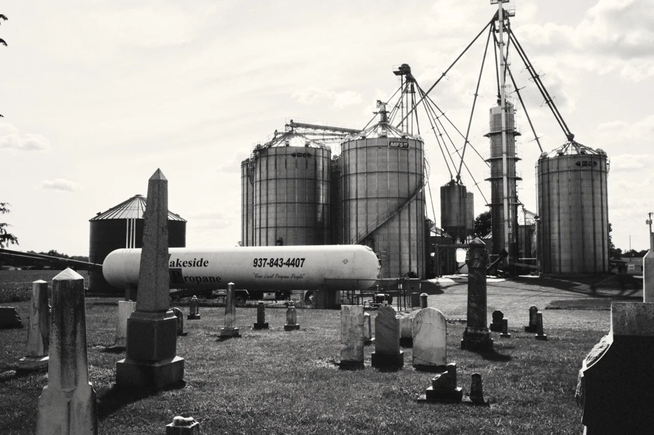 Graveyard with headstones in the foreground and silos and industrial structures in the background, depicted in black and white.