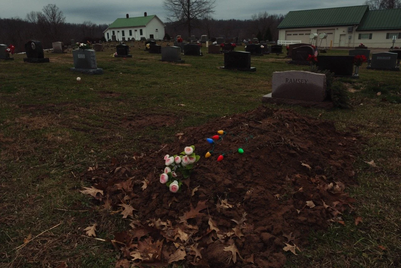 Grave site with a mound of earth, pink roses, and colorful plastic Easter eggs, in a cemetery with multiple headstones and a white building with a green roof in the background.