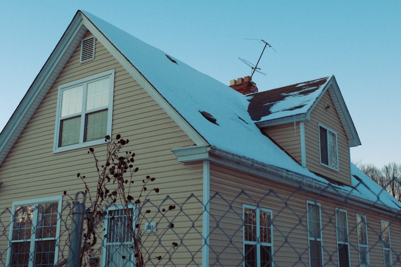 Yellow house with snow on the roof, behind a chain-link fence, with multiple windows and a leafless shrub in front.