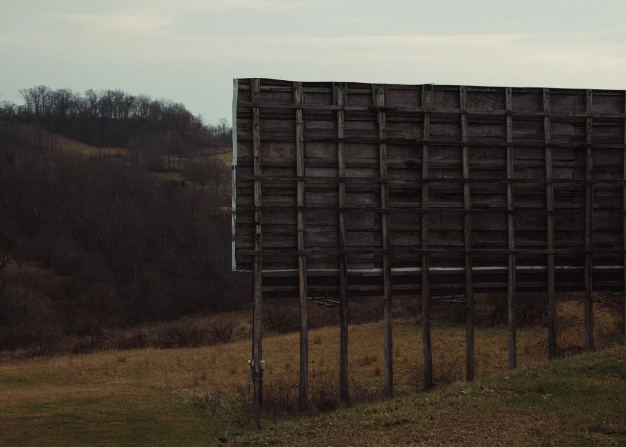 A weathered wooden billboard structure in a rural area with bare trees and hills in the background.