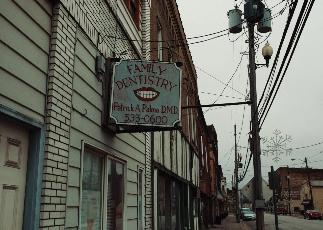 Street view of a small town with a dental office sign reading "Family Dentistry" by Patrick A. Palma DMD, with a phone number, along with buildings, utility poles, and snowflake holiday decorations hanging from wires.