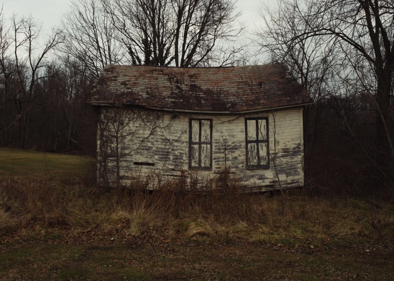 An old, dilapidated house with peeling white paint, broken windows, and an overgrown yard, surrounded by leafless trees on a cloudy day.