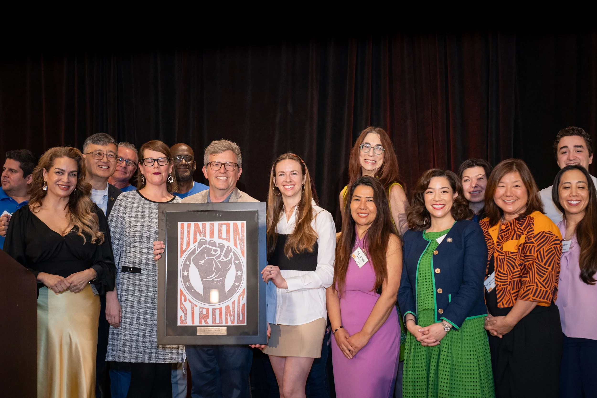 A group of diverse people standing together on stage, smiling, with one person holding a framed sign that says 'UNION' and features a raised fist.