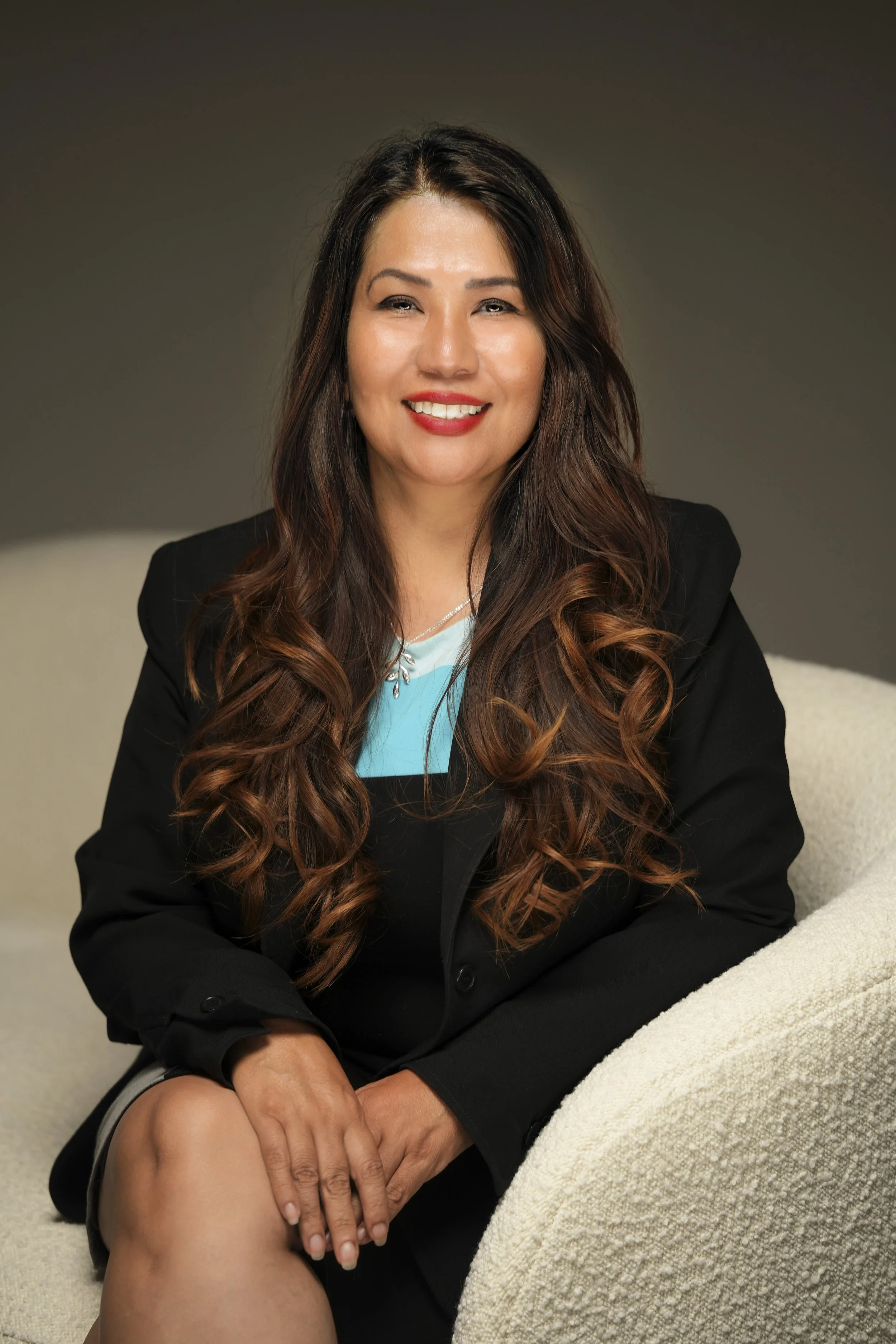 A smiling woman with long wavy brown hair, wearing a black blazer and a blue top, sitting on a cream-colored couch.
