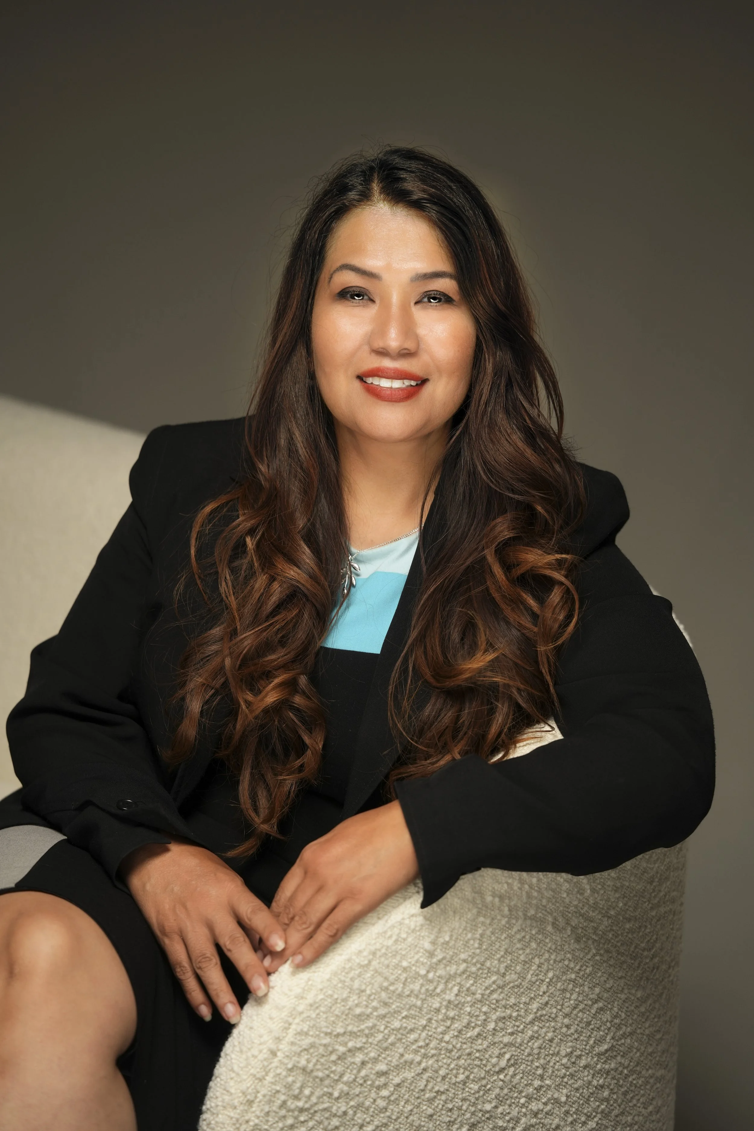 A woman with long, wavy brown hair, wearing a black blazer over a blue top, smiling softly and sitting on a gray couch with a neutral-colored background.