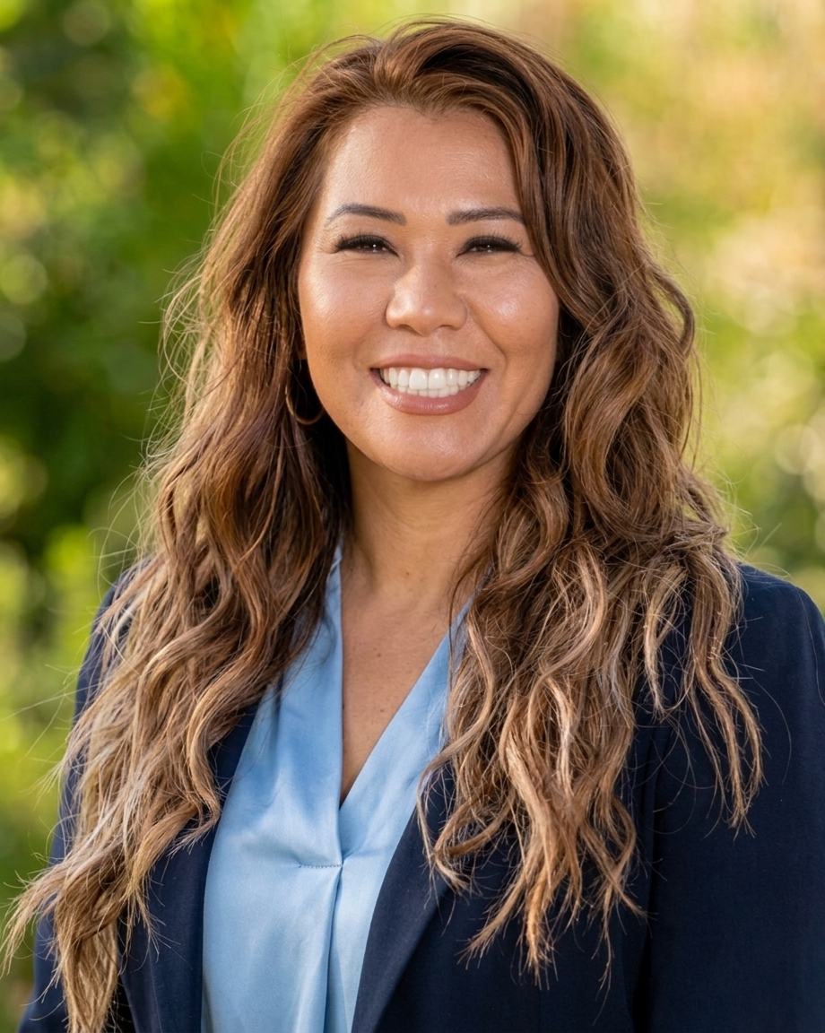 A smiling woman outdoors with long wavy brown hair, wearing a light blue blouse and dark blazer, with a blurred green background.