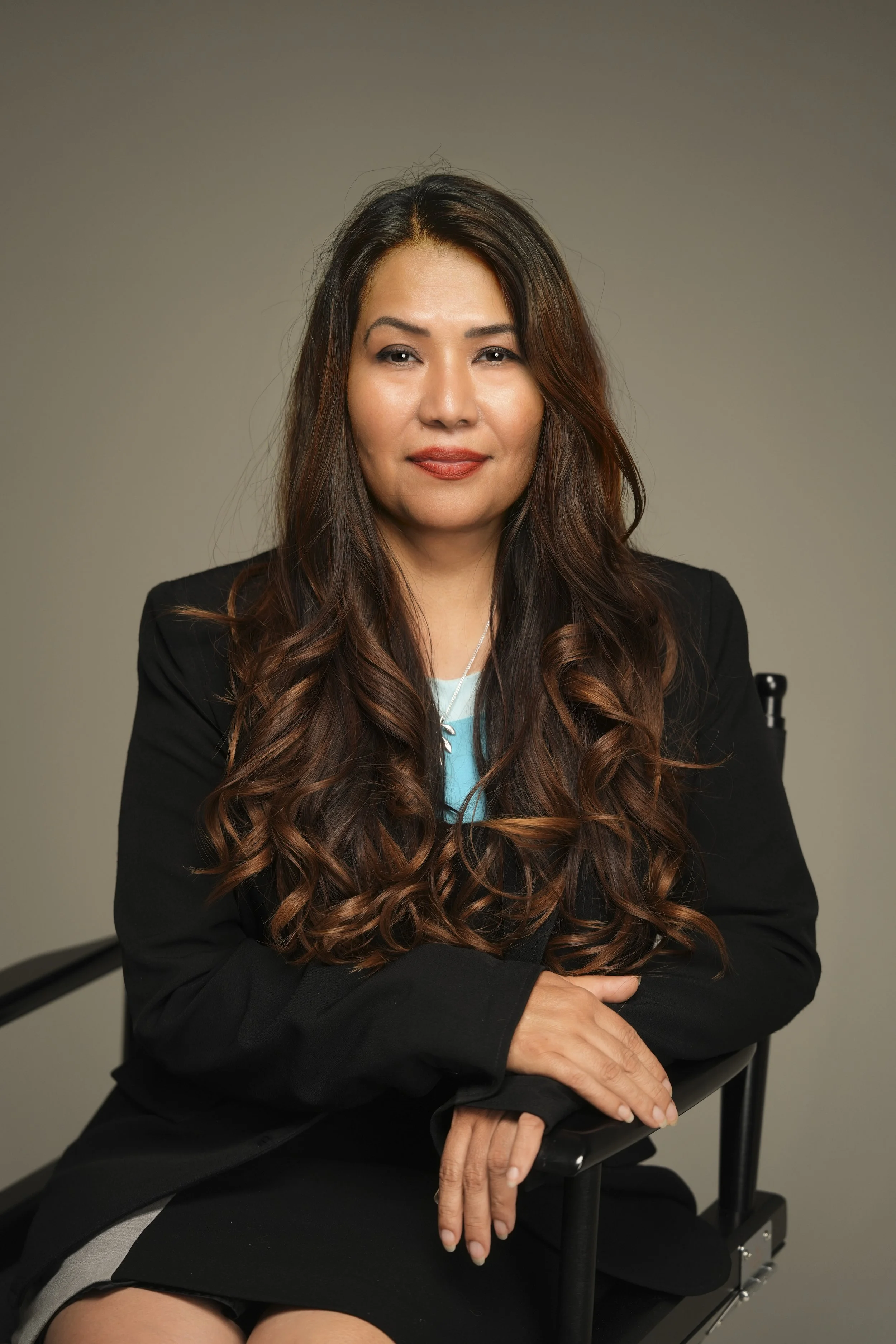 A woman with long wavy brown hair, tan skin, and wearing a black blazer, sits with her hands crossed on her lap, on a black chair against a plain beige background.