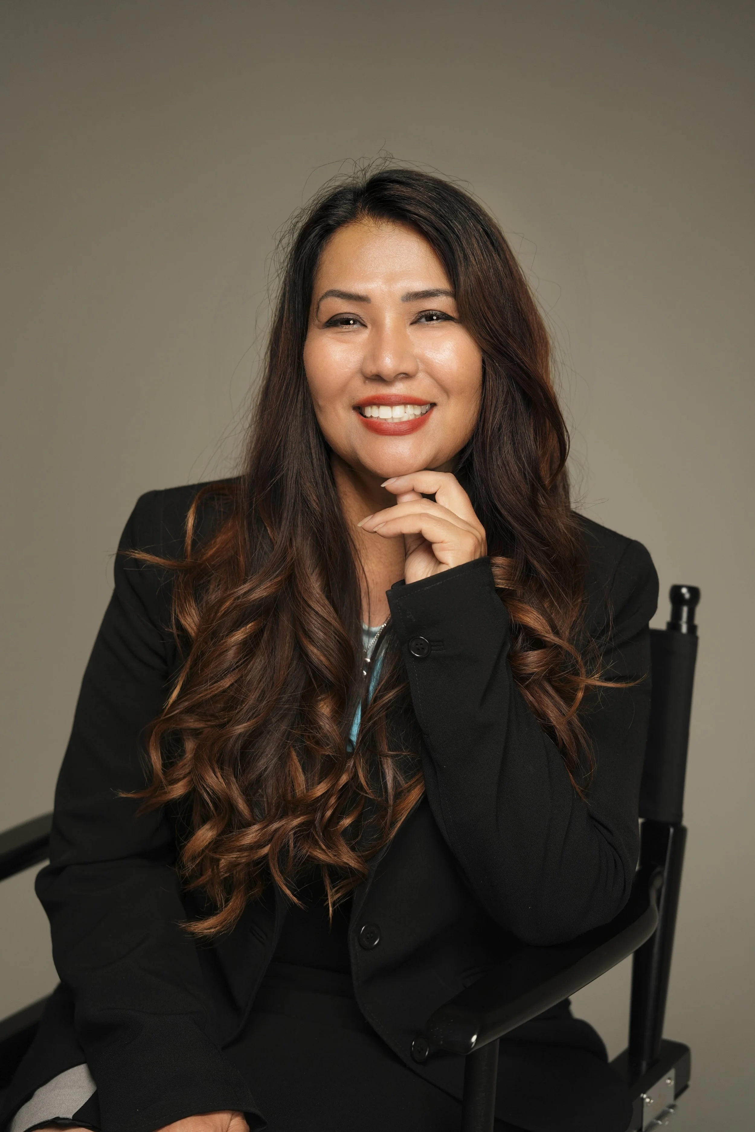 A woman with long, wavy brown hair smiling, sitting on a black chair, wearing a black blazer, against a neutral background.
