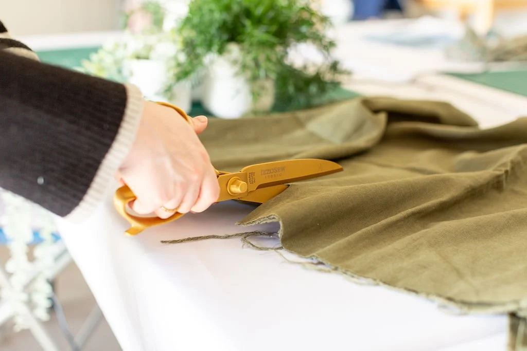 A close-up of hands cutting though green linen fabric using a pair of gold tailors scissors.