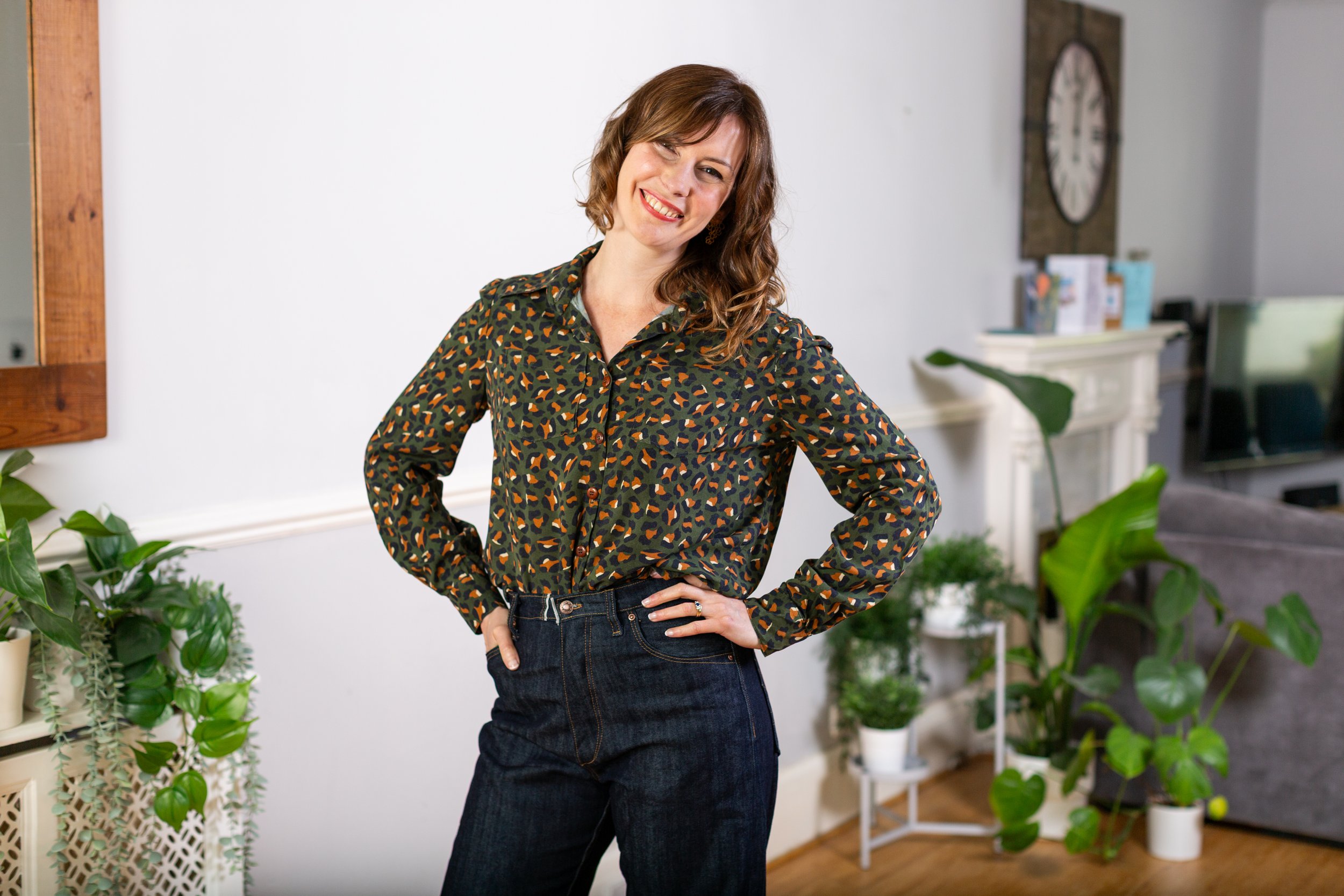 The sewing tutor smiling warmly while wearing a self-drafted leopard print shirt and dark denim trousers, standing in an inviting, plant-filled home studio.