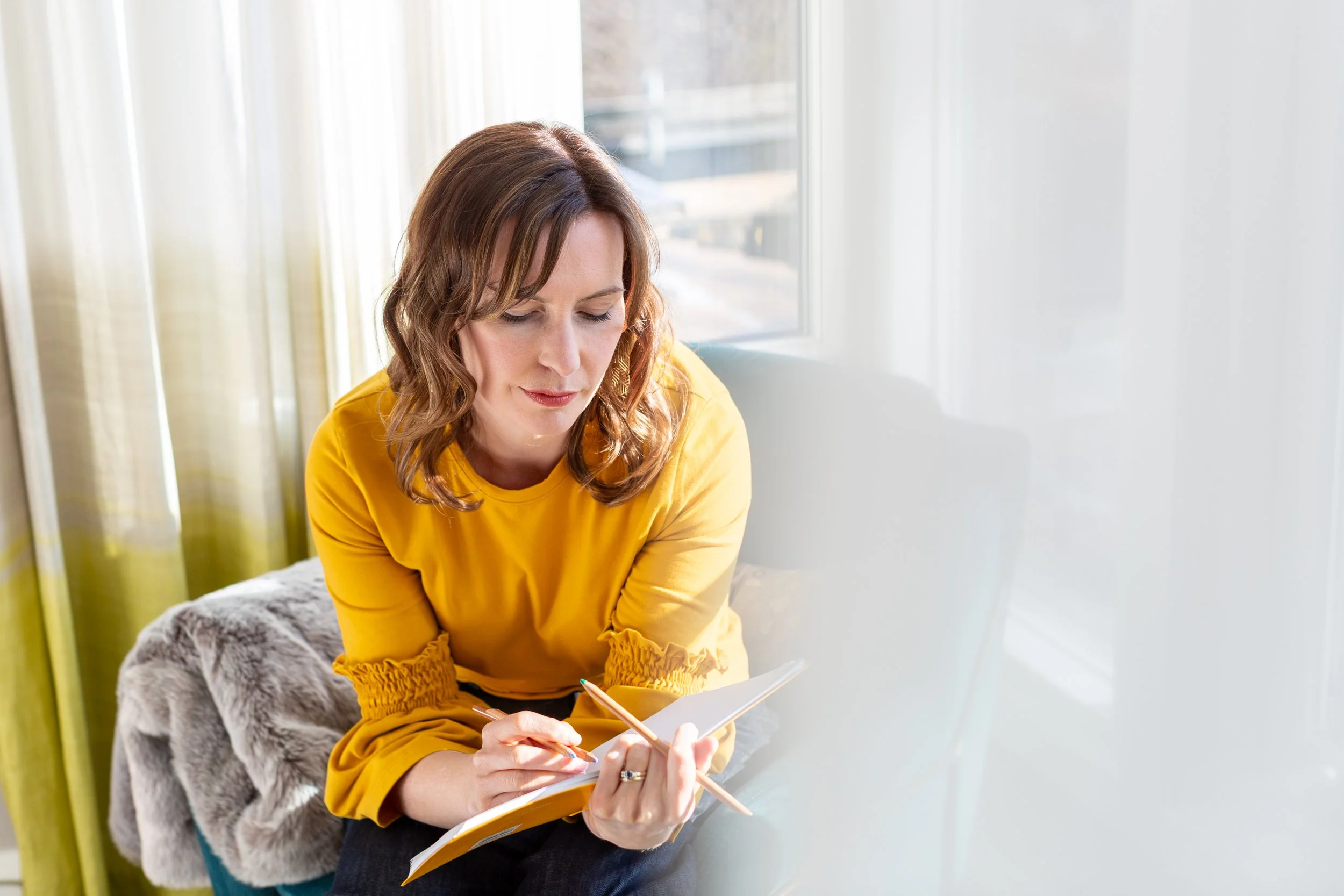 A person sitting in a relaxed lounge environment, sketching her garment designs in a sketch book, looking thoughtful.