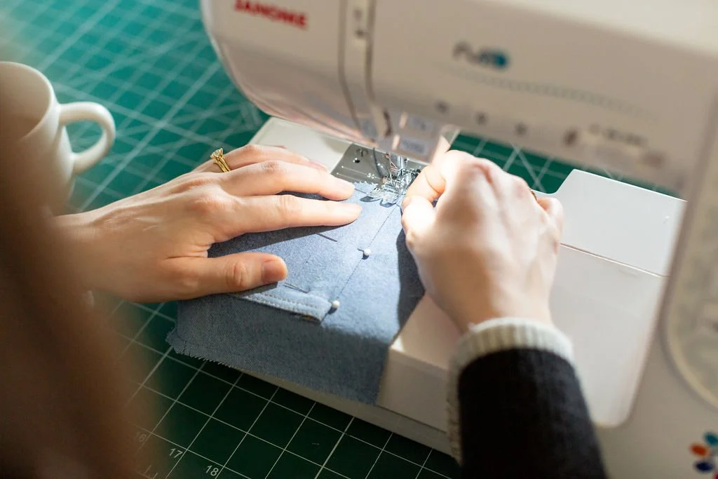 A close-up of a maker carefully guiding blue denim fabric through a sewing machine, topstitching a pocket detail.