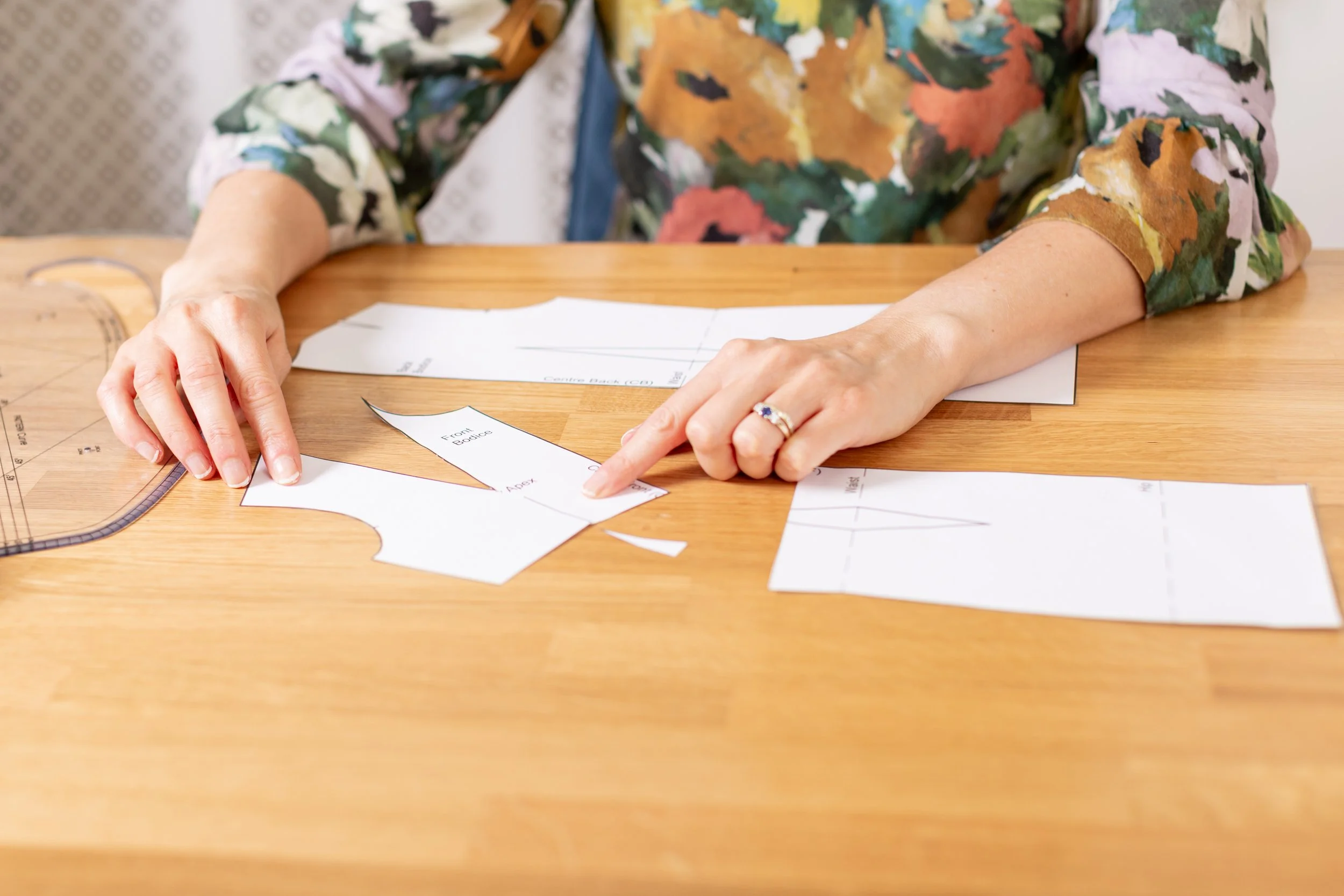 A close-up of hands performing technical alterations on a paper dress block pattern, featuring a dart manipulation and a French curve tool on a wooden workspace.