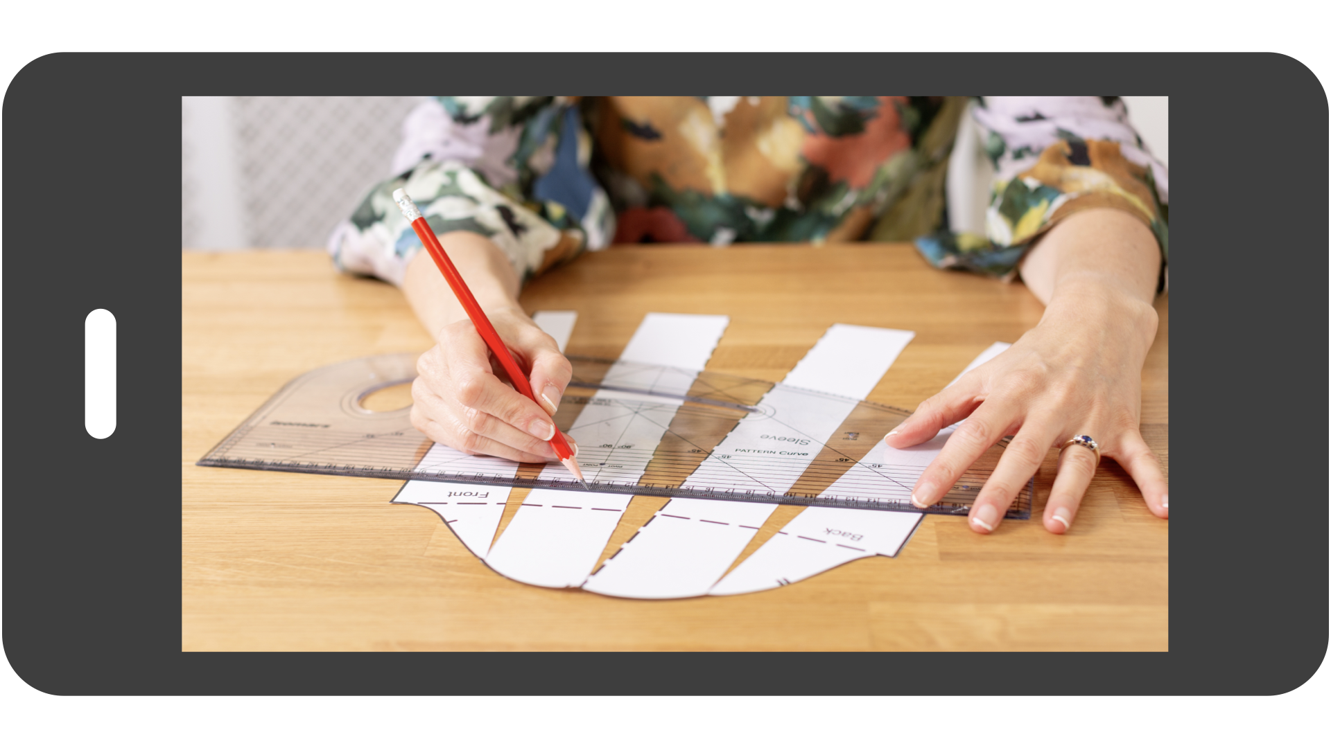 A digital smartphone preview of a technical sewing lesson, showing a close-up of hands using a red pencil and professional grading ruler to perform a slash-and-spread adjustment on a paper sleeve pattern.