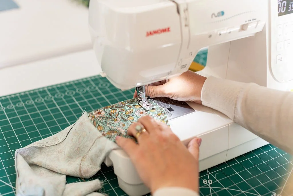 A close-up of hands with manicured nails and a gold ring carefully guiding a green floral fabric under the presser foot of a white Janome sewing machine, demonstrating professional garment construction.