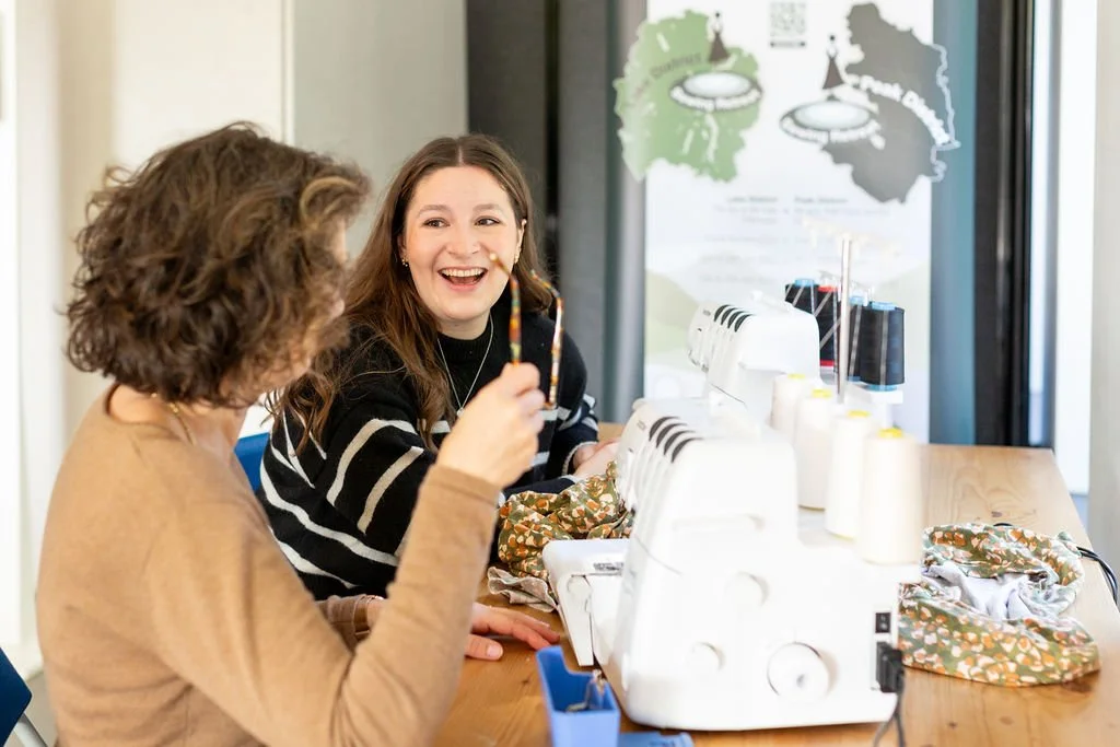 Two women laughing and chatting while working with white overlocker machines at a sewing workshop.