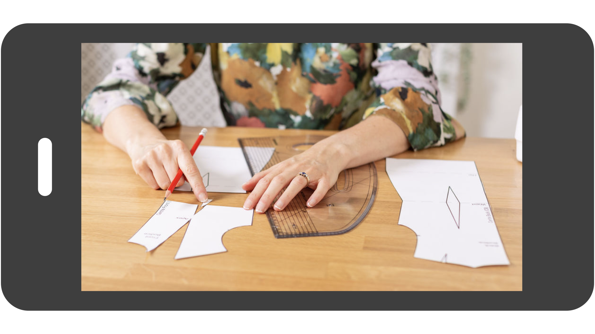 A digital smartphone preview of a professional pattern drafting tutorial, showing a close-up of hands using a red pencil to demonstrate dart manipulation on a slashed paper bodice block next to a clear French curve ruler.