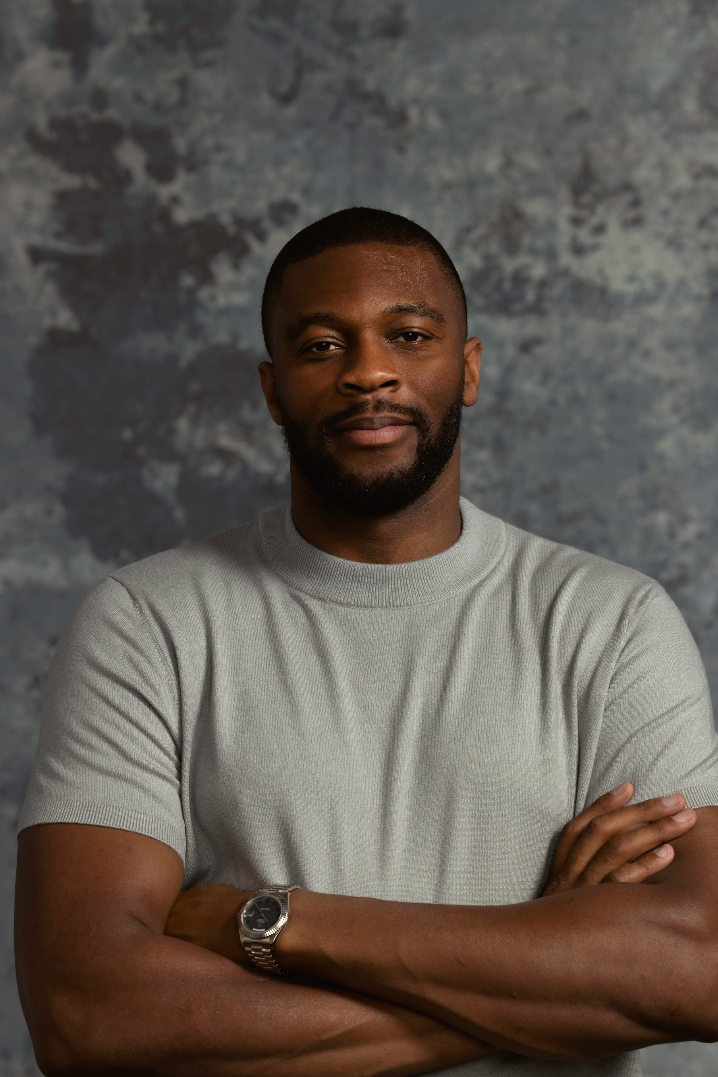 Portrait of a confident man with crossed arms, wearing a light gray t-shirt and a silver watch, standing against a textured gray background.