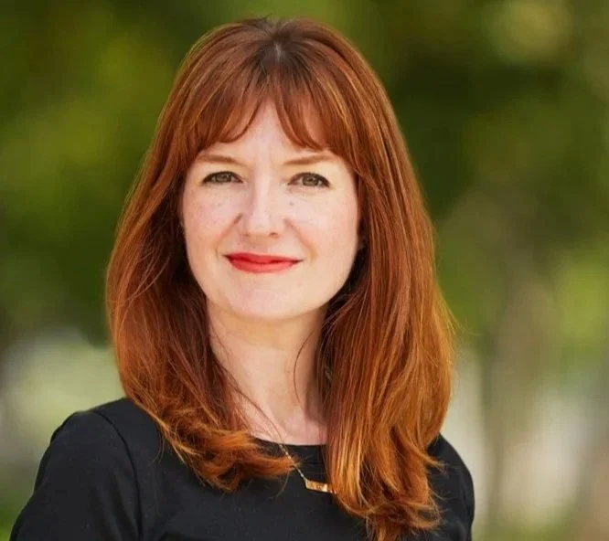 A woman with red hair and fair skin smiles outdoors with a blurred green background.